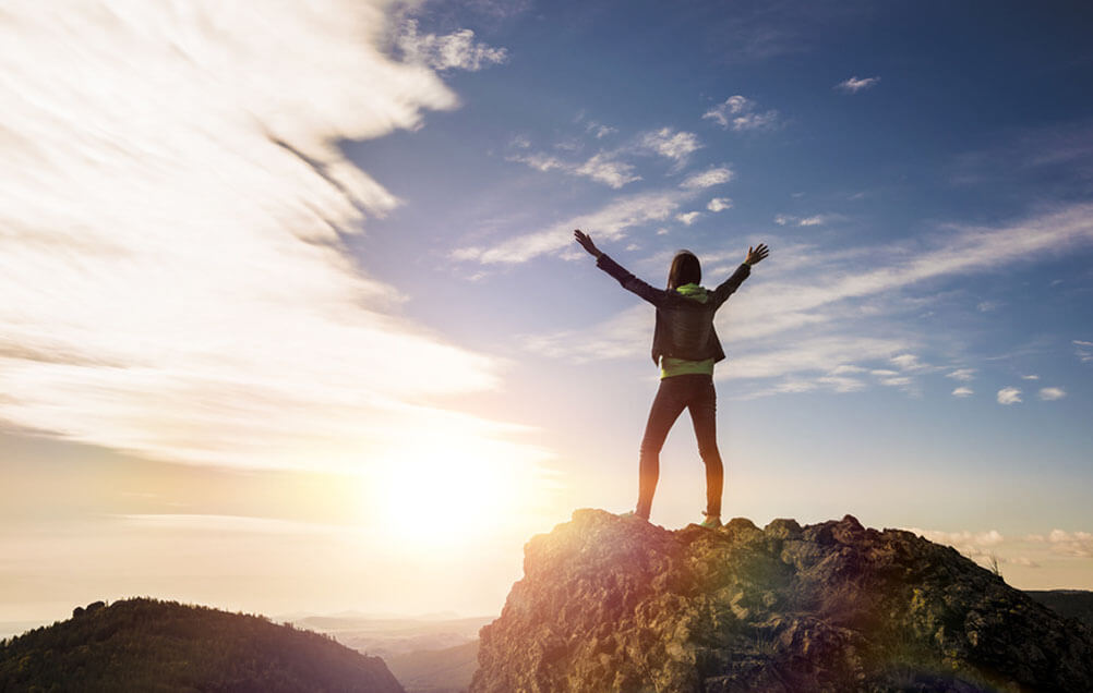 Woman stands on top of a mountain with arms outstretched as the sun sets